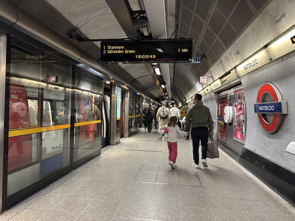 U-Bahn-Station mit Zügen an den Gleisen und Passagieren, die auf den Bahnsteig gehen, während eine Anzeige die nächsten Abfahrten zeigt in London.