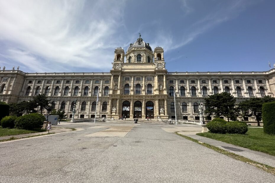 Historisches Gebäude mit beeindruckender Architektur, das eine zentrale Fassade und große Eingänge aufweist, umgeben von gepflegten Grünflächen.