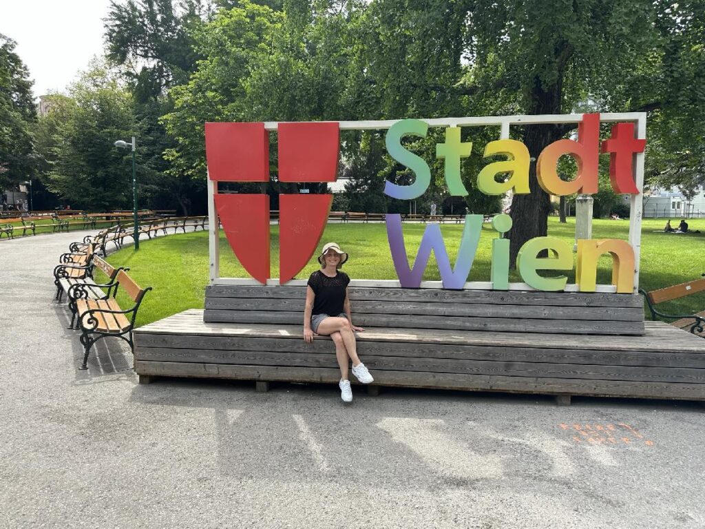 Frau sitzt auf einer Bank vor einem bunten Schild mit der Aufschrift 'Stadt Wien' in einem Park.
