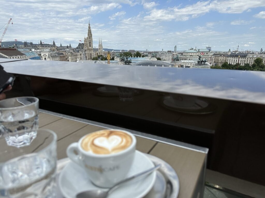 Tasse mit Cappuccino und Herzmuster auf einem Tisch, im Hintergrund eine Stadtansicht mit historischen Gebäuden und Himmel.