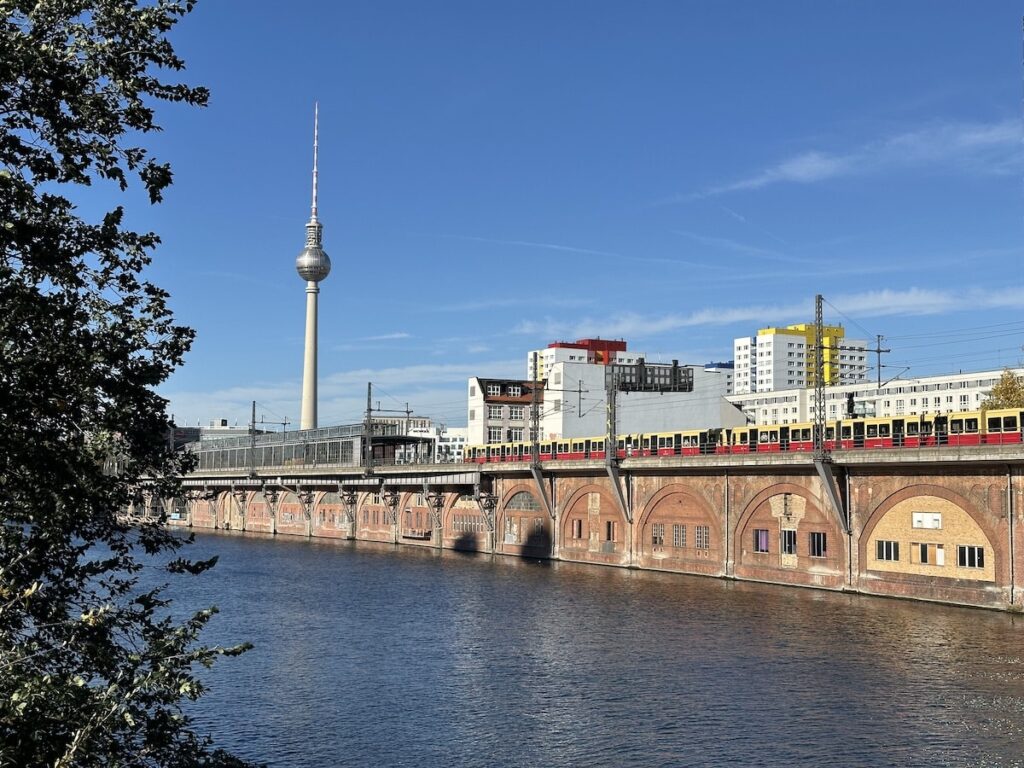 TV tower in Berlin with an S-Bahn train running along the Spree River, water and riverbank vegetation in the foreground.