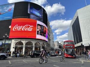 Cyclists and a red double-decker bus pass beneath giant Coca-Cola and Virgin Atlantic adverts at Piccadilly Circus in London.