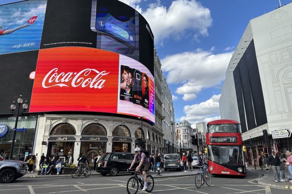 Großes Werbebildschirm mit Coca-Cola-Logo und weiteren Anzeigen in einer belebten Stadtstraße mit Fahrrädern und roten Doppeldeckerbussen.