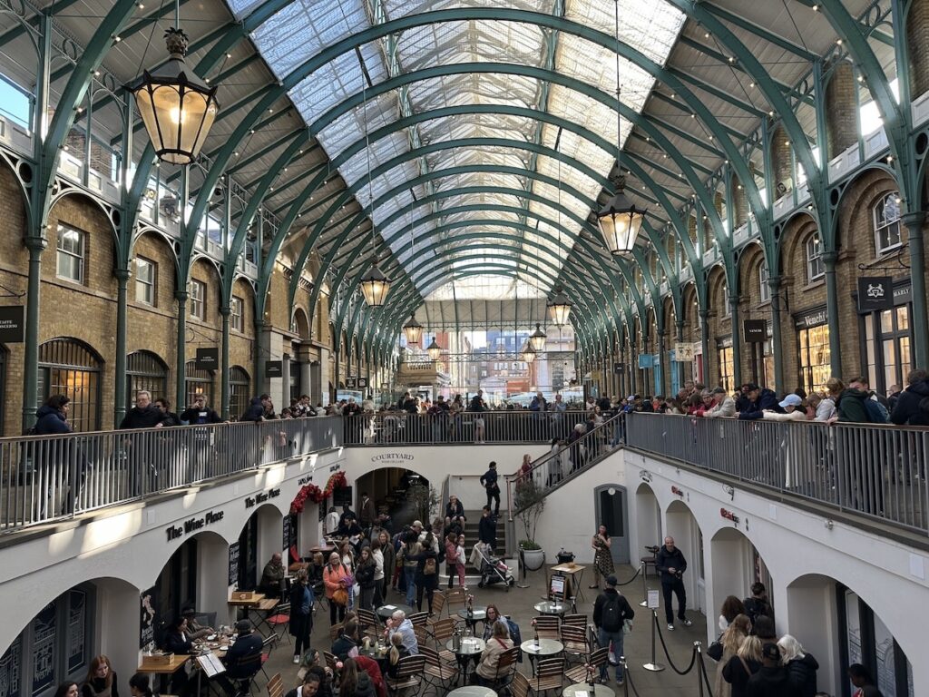 Interior view of a market hall with a high glass roof and many people moving around on several levels.