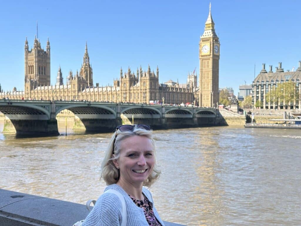 Frau mit blonden Haaren steht am Ufer der Themse mit dem Big Ben und dem Palace of Westminster im Hintergrund.