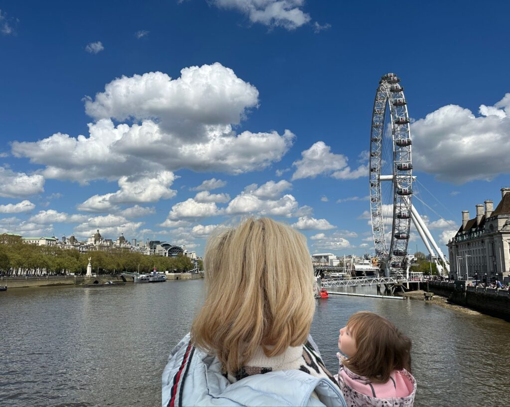 Woman with blonde hair and child in her arms, looking at the London Eye and the sky with clouds.