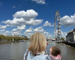 Woman with blonde hair and child in her arms, looking at the London Eye and the sky with clouds.
