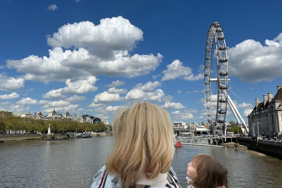 Woman with blonde hair and child in her arms, looking at the London Eye and the sky with clouds.