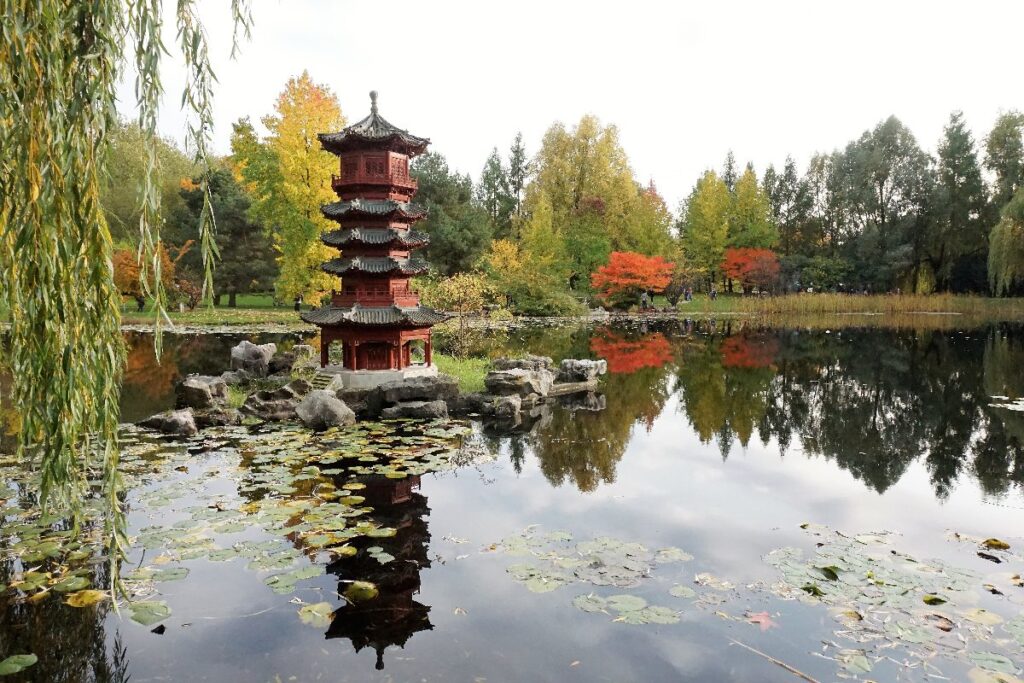 A red Japanese-style pagoda stands beside a pond in a Berlin park, with autumn trees and lily pads reflected in the water.