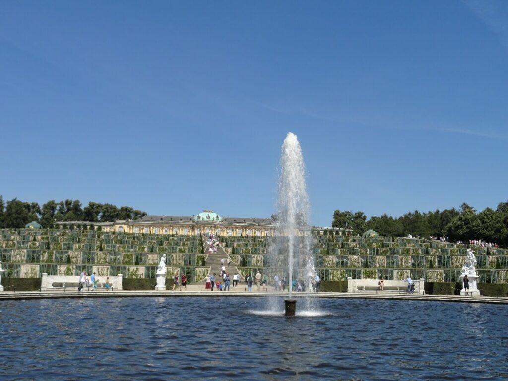 Large fountain in the terraced gardens of Sanssouci Palace in Potsdam, with visitors walking towards the palace on a sunny day.
