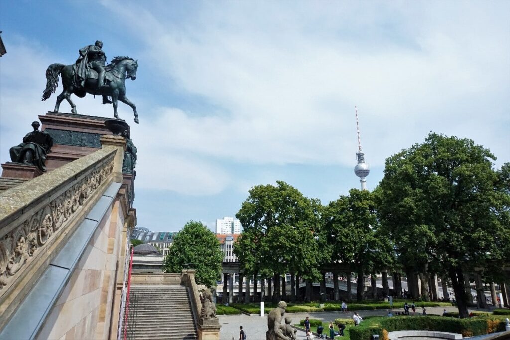An equestrian statue rises above Museum Island’s stone steps, with leafy trees and the Berlin TV Tower visible beyond.
