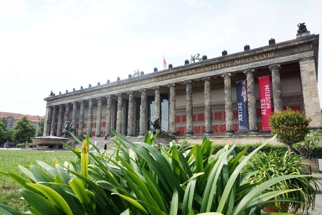 A grand columned Altes Museum stands on Berlin’s Museum Island, its red banners rising above leafy garden plants.
