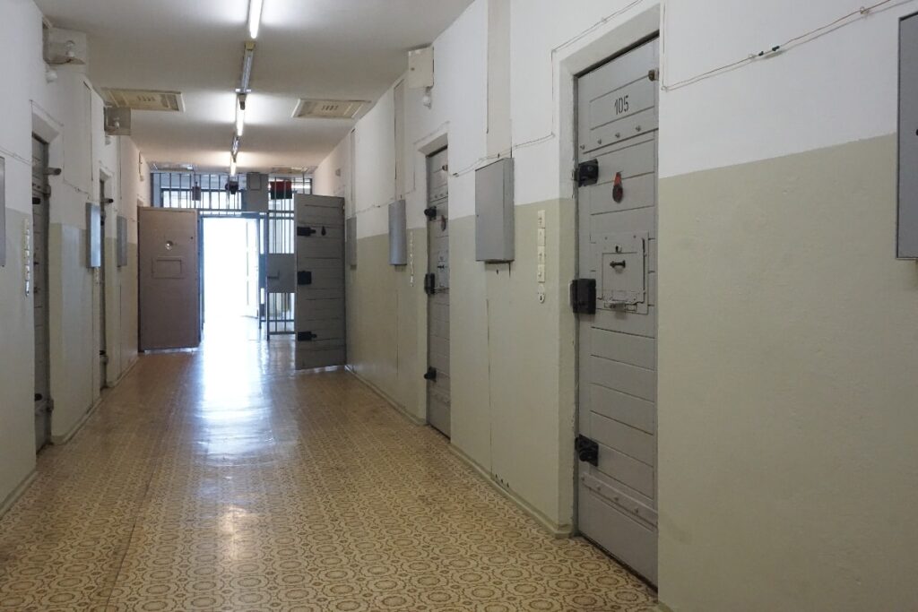 A stark prison corridor in Berlin has heavy cell doors lining the passage and a barred gate at its far end.