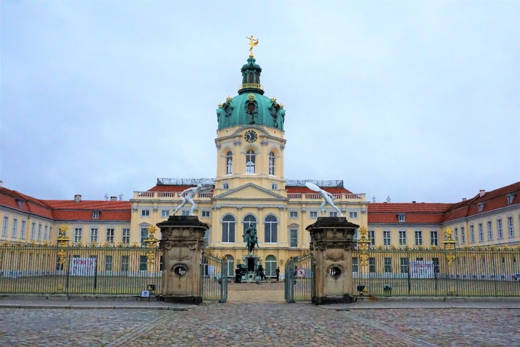 The yellow Charlottenburg Palace in Berlin rises behind ornate gates, with a clock tower crowned by a green dome.