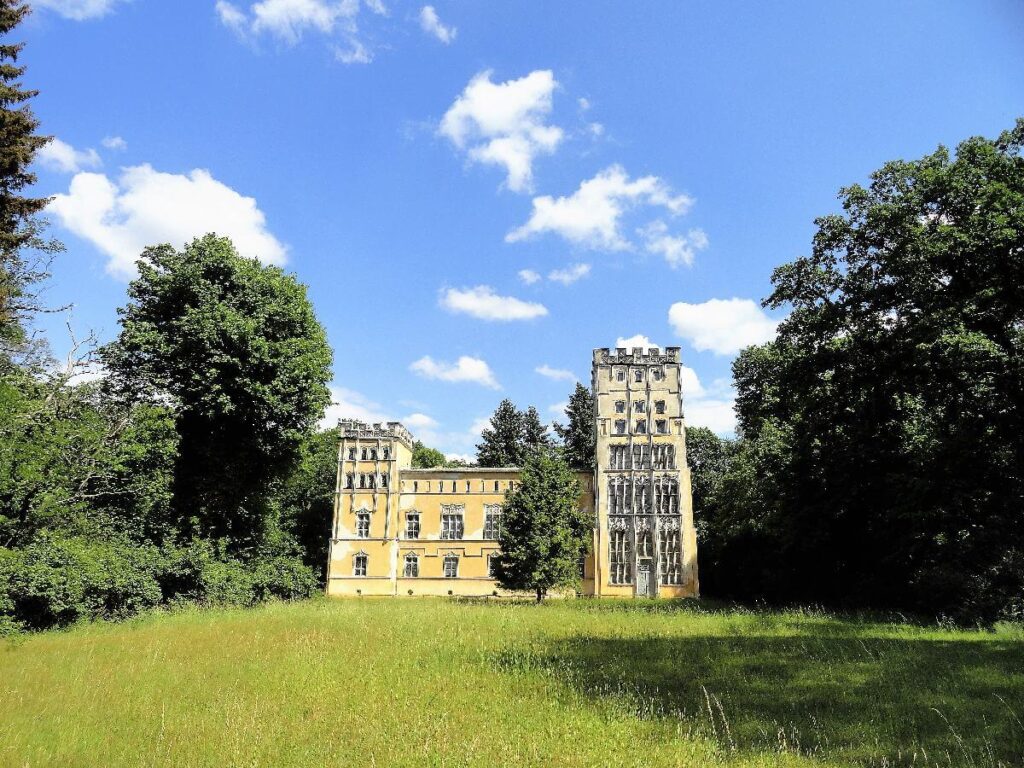 A historic yellow building with ornate towers stands amid leafy trees and a grassy meadow beneath a bright blue sky.