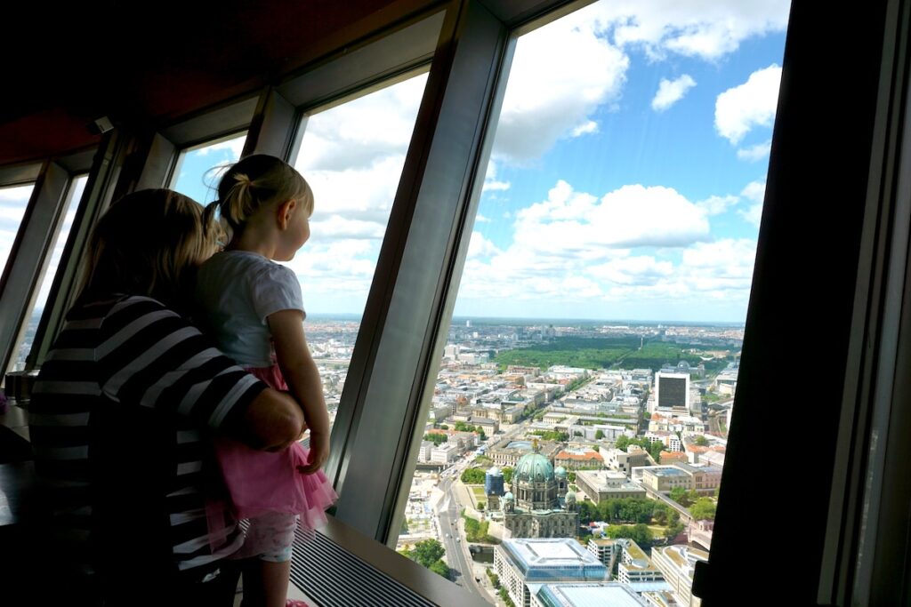A mother and daughter look out from Berlin’s TV Tower observation deck at the city skyline, with the Berlin Cathedral below.