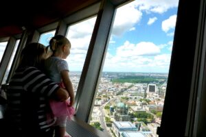 A mother and daughter look out from Berlin’s TV Tower observation deck at the city skyline, with the Berlin Cathedral below.