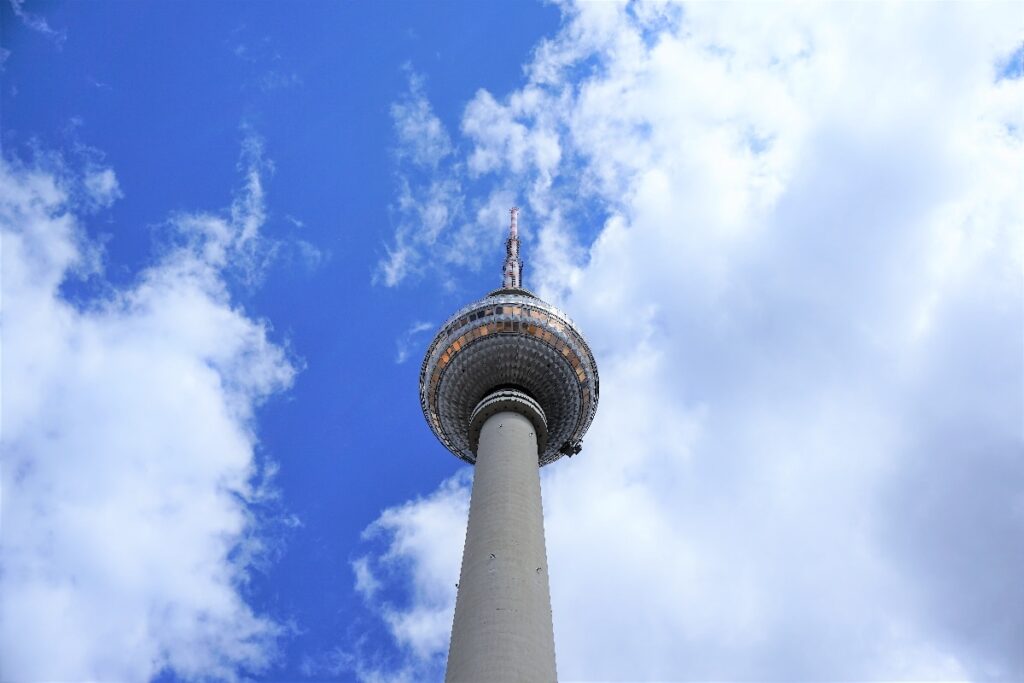 Looking up at Berlin TV Tower, its observation sphere and tall concrete shaft rise against a vivid blue sky filled with clouds.