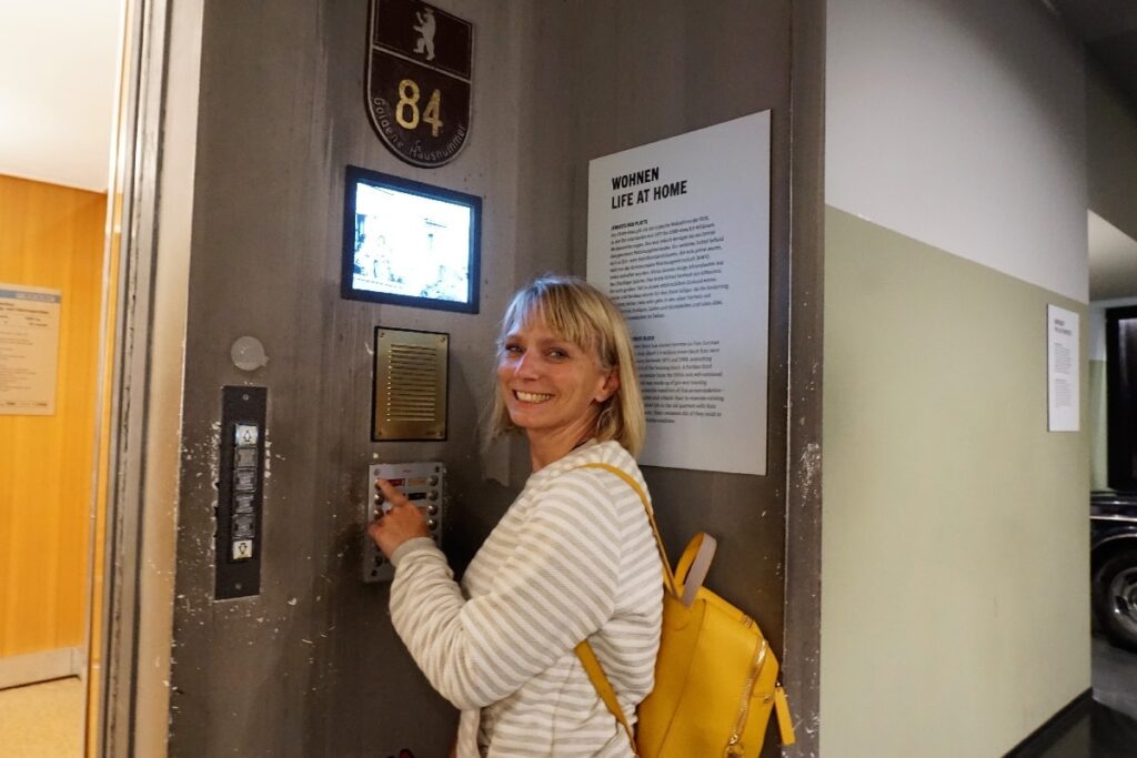 A smiling visitor presses an interactive museum button beneath a display about life at home, exploring indoor activities in Berlin.