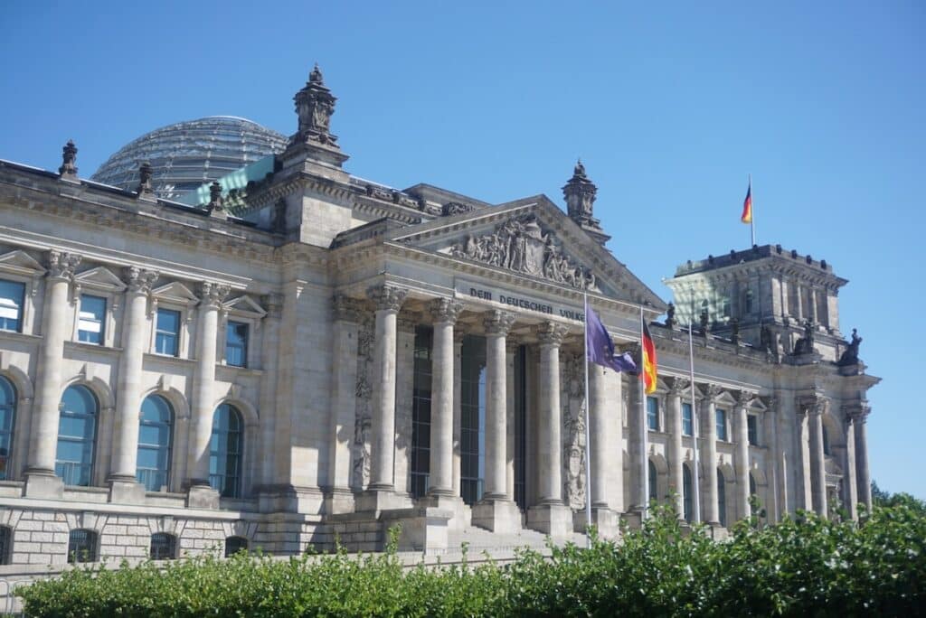 The Reichstag building in Berlin with its glass dome, historic stone facade and German flags under a clear blue sky.