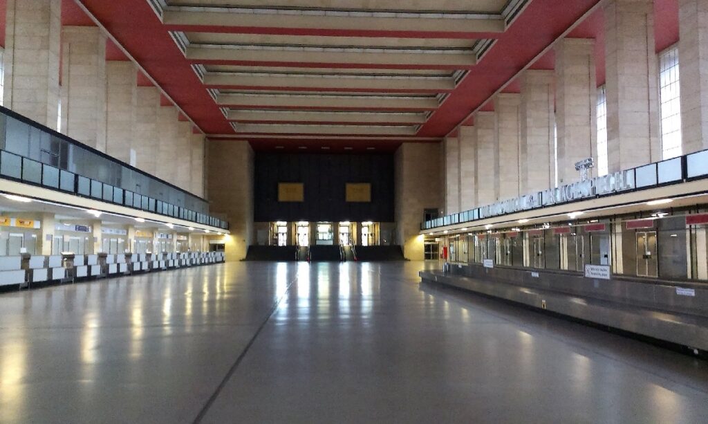 An empty Berlin indoor hall has long service counters along both sides, beneath a high red ceiling and tall pale stone walls.