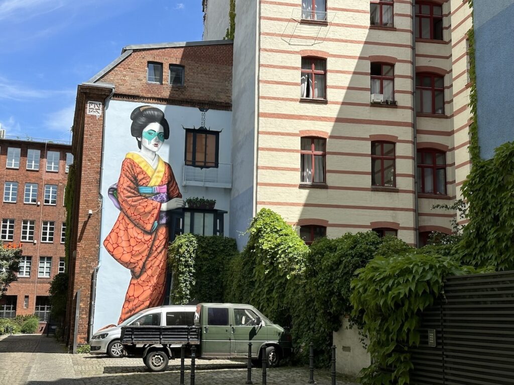Large mural of a Japanese geisha in an orange kimono on a building wall in a Berlin courtyard.