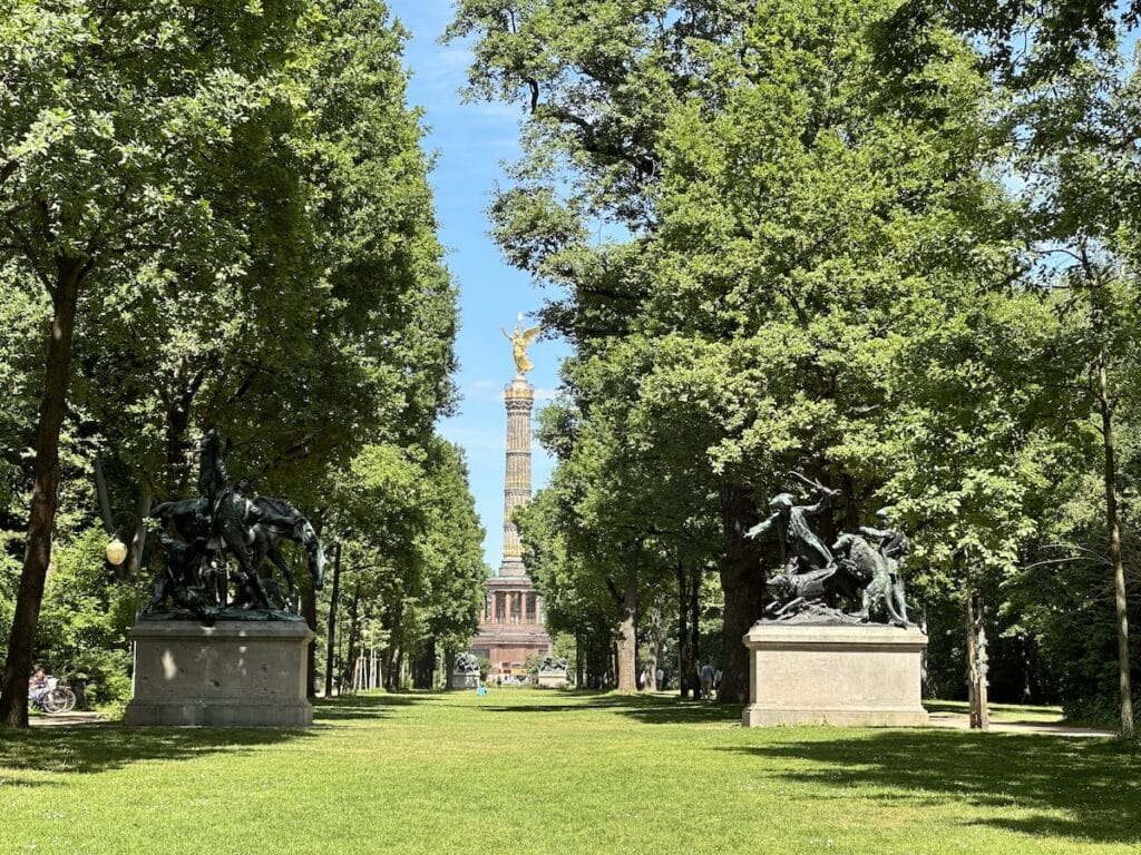View of the Victory Column (Siegessäule) in Berlin's Tiergarten park, framed by green trees and historic statues.