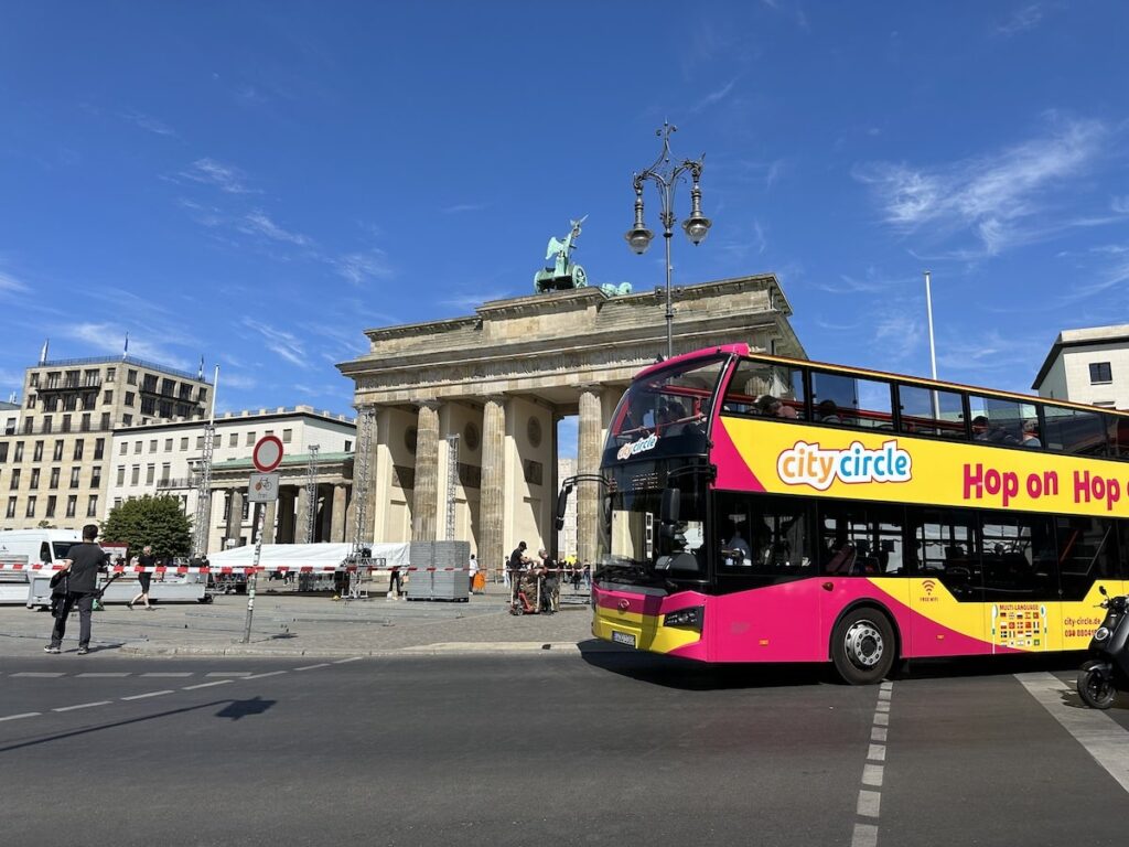 Pink and yellow City Circle hop-on hop-off sightseeing bus in front of the Brandenburg Gate in Berlin on a sunny day.