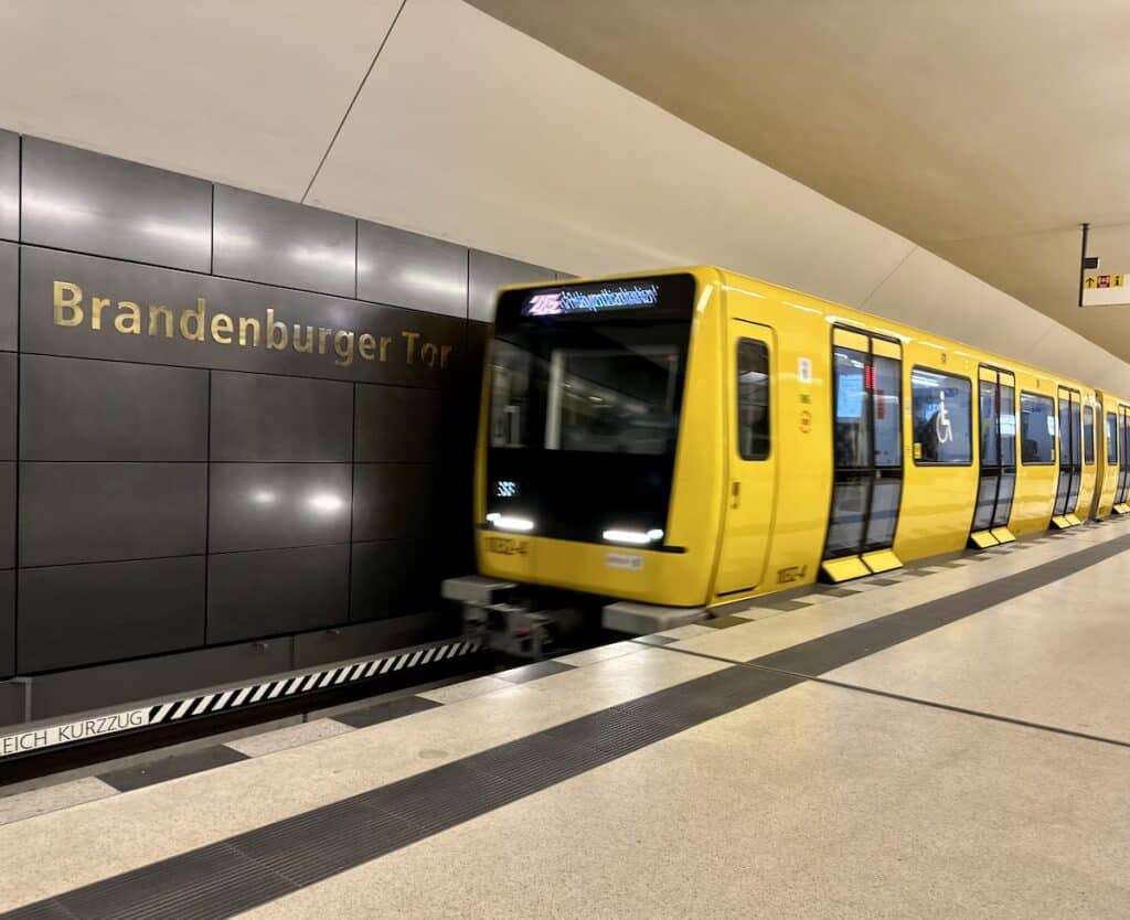 A yellow Berlin U-Bahn train passes the Brandenburger Tor station sign on its way through the underground platform.