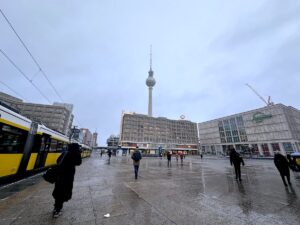 Rain-soaked Alexanderplatz in Berlin, with a yellow tram passing beneath the Fernsehturm as pedestrians cross the square.