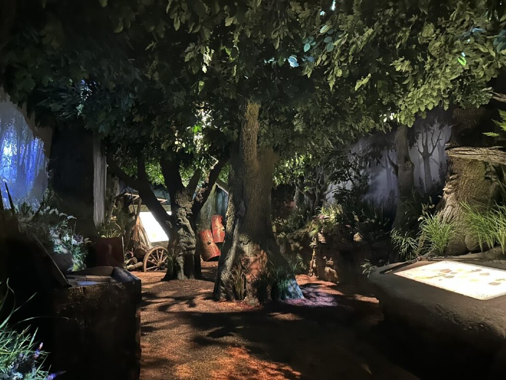 A dimly lit forest setting at the Deutschlandmuseum features towering trees, a rustic wagon, plants and interactive display panels.