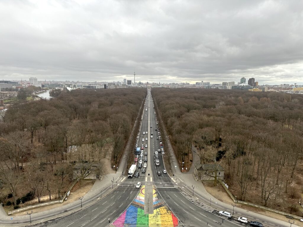 A busy avenue crosses a leafless Berlin park toward the Fernsehturm, with a colorful rainbow crossing beneath heavy clouds.