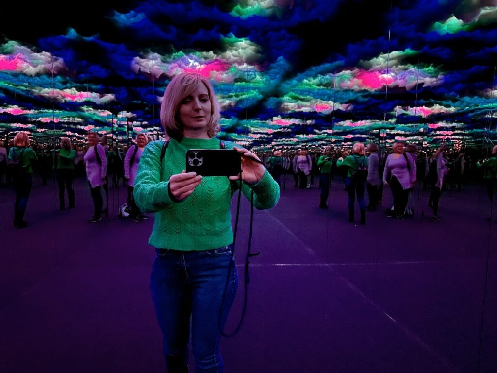 A woman photographs herself among mirrored reflections and colorful cloud-like lights at the Illuseum, an indoor activity in rainy Berlin.