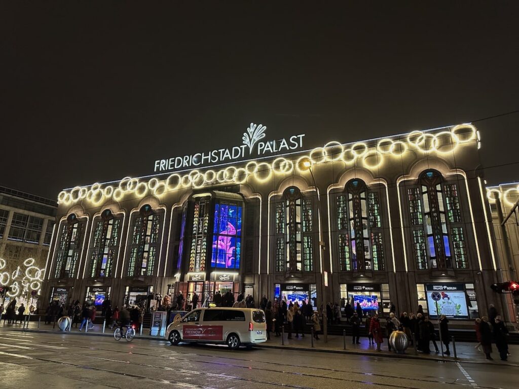 At night, people gather outside Berlin’s Friedrichstadt-Palast, where circular lights frame the theater’s illuminated sign and facade.
