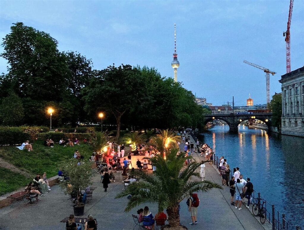 People gather along the River Spree at dusk in Berlin, with the illuminated TV Tower rising above the trees.