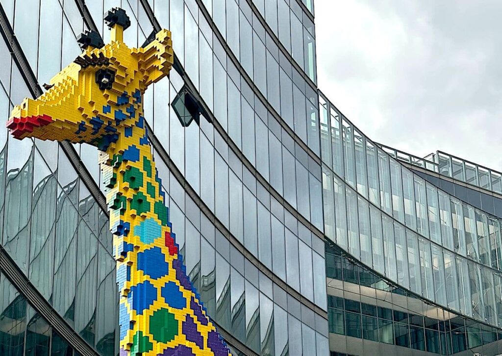A pixel-style giraffe sculpture with a raised head stands beside a glass-fronted building, adding a playful note to rainy-day Berlin.